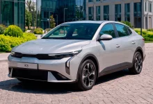 Серебристый седан Kia K3 спереди сбоку на брусчатке у стеклянного офисного здания
