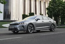 Серый седан Kia K5 спереди сбоку на дороге у здания с белыми колоннами