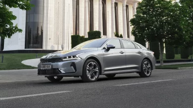 Серый седан Kia K5 спереди сбоку на дороге у здания с белыми колоннами