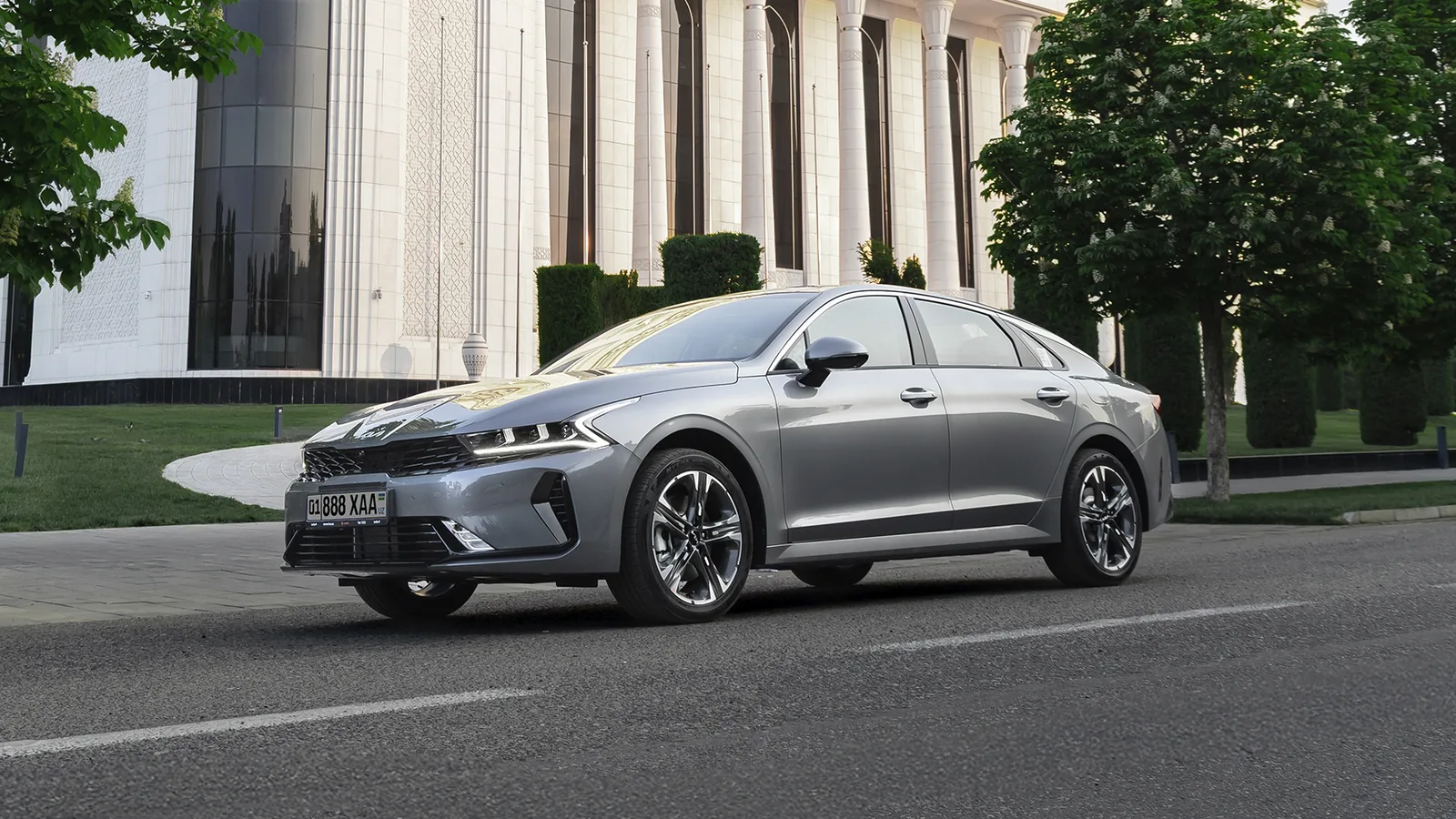 Kia K5 в рассрочку без процентов: взнос и ежемесячный платеж - 1 Серый седан Kia K5 спереди сбоку на дороге у здания с белыми колоннами